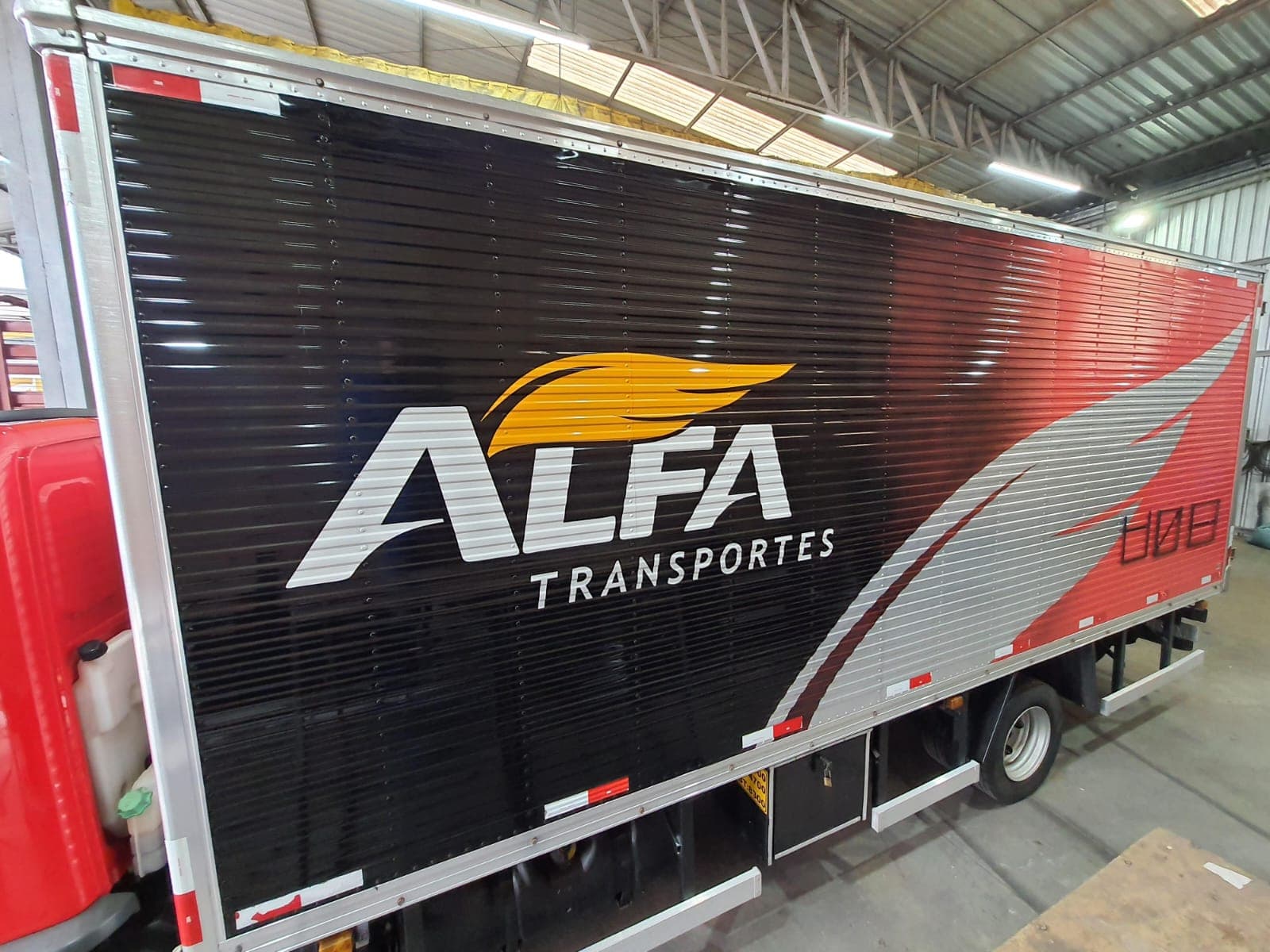 Caminhão baú da Alfa Transportes com a logomarca em branco e amarelo sobre o preto e grafismo vermelho e prata na lateral